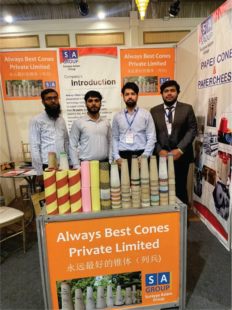 A promotional booth for 'Always Best Cones Private Limited' showing a group of four individuals with paper cones stacked in front of a bright orange display banner.