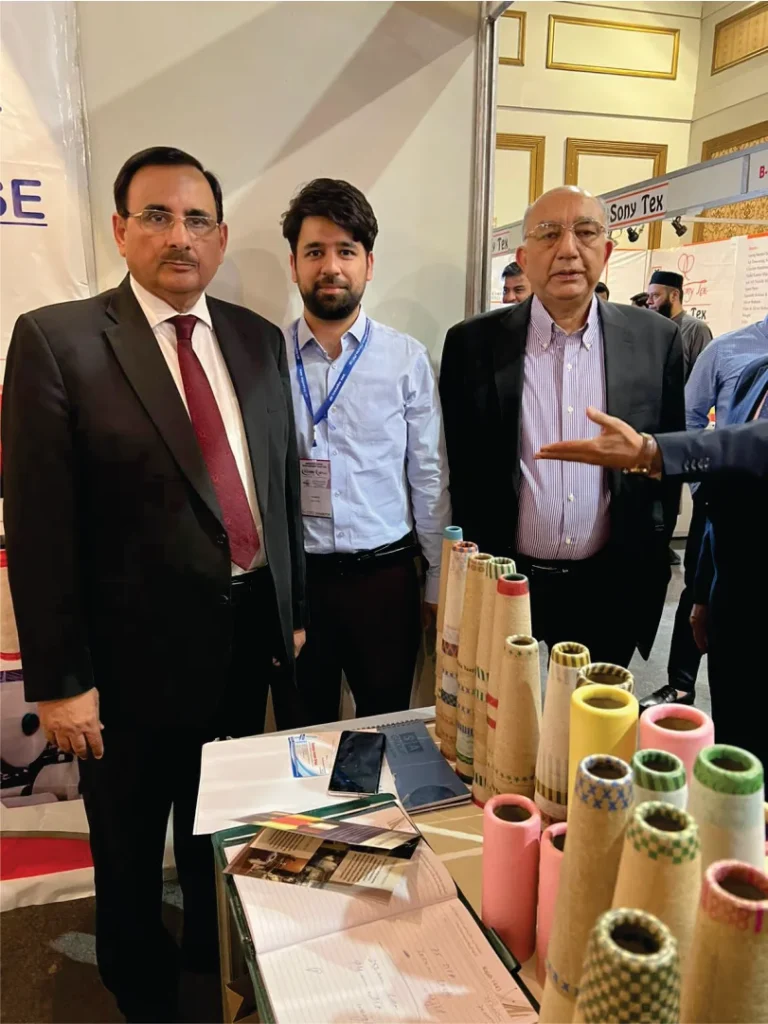 Two men discussing a sample of paper cones at a booth, with other attendees and a colorful display in the background.